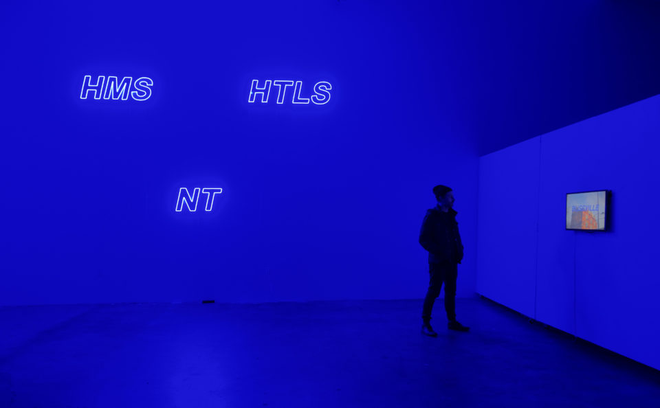 Image shows blue neon letters HMS NT HTLS and person looking at a video screen withe the world BLISSVILE in front of a building